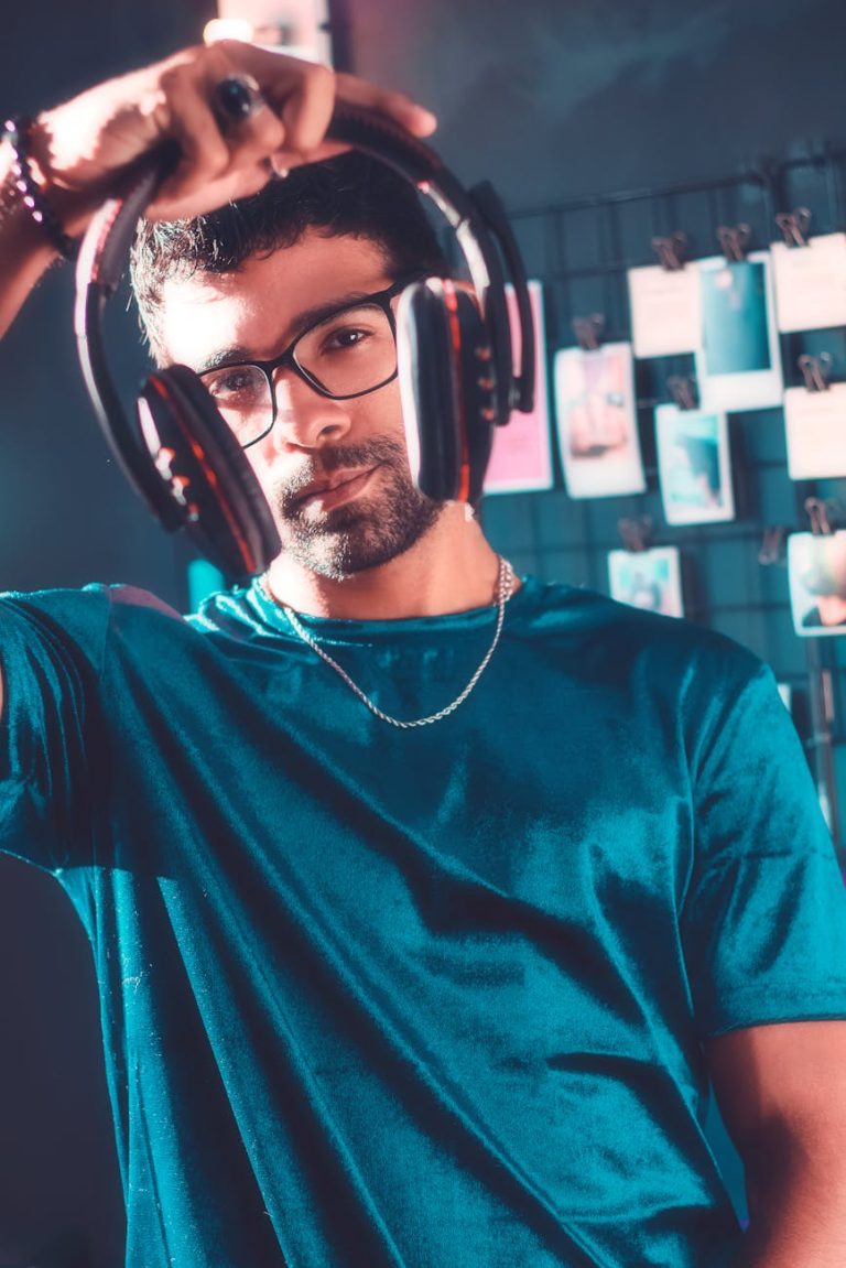 Fashionable man with glasses holding headphones, wearing a t-shirt in a modern indoor setting.