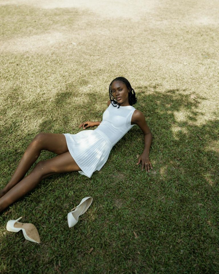 A stylish woman in a white dress relaxes on a sunlit grass field with shoes off, enjoying a peaceful day outdoors.