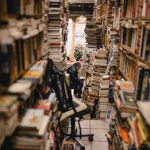 A narrow aisle filled with stacks of books in a cozy, crowded bookstore, featuring a person reading.