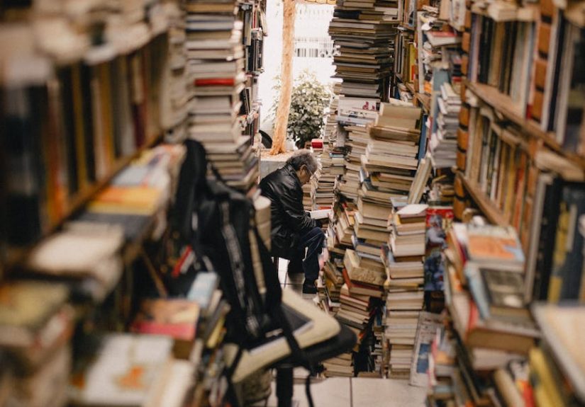A narrow aisle filled with stacks of books in a cozy, crowded bookstore, featuring a person reading.